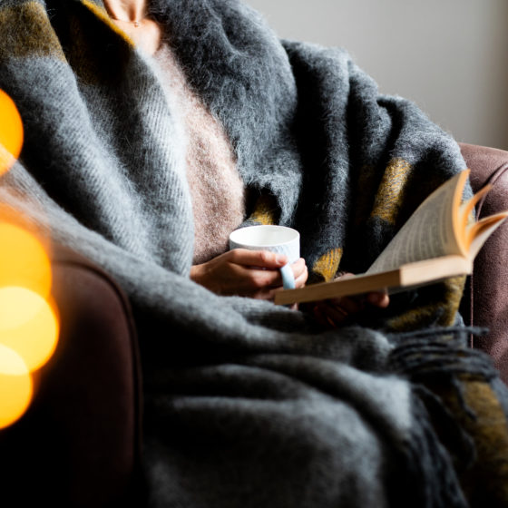 Une personne enroulée dans une couverture lit un livre et boit du café.