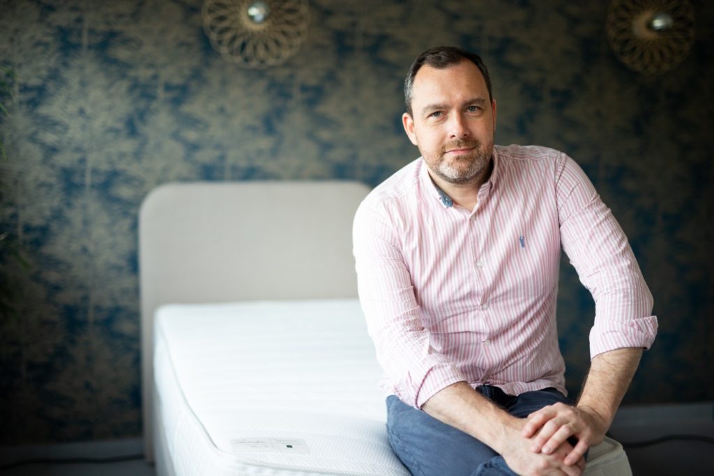 Un homme assis sur un lit, chemise rose à rayures.