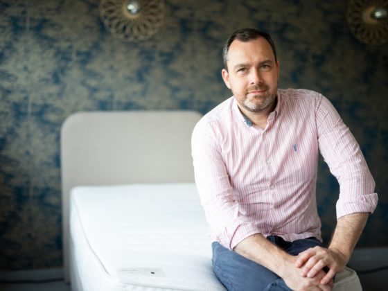 Un homme assis sur un lit, chemise rose à rayures.